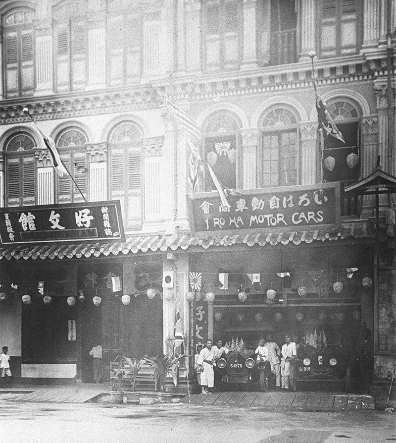 Kobunkan bookstore on the left and Iroha Motor Cars & Co. on the right at 167 and 168 Middle Road respectively, c. 1910s. The handwritten caption behind the photograph reads 「英領新架坡中央街　山﨑・竹井経営　日本人初の自動車営業　日本人初の書籍新聞販売店　西宇和郡（現西予市）三瓶町出身竹井庫敬（くらよし＝向かって左の和服の男性）ほか馬来人、車掌、ボーイ」[Eiryō Shingapōru Chūō-gai Yamazaki・Takei keiei Nihonjin hatsu no jidōsha eigyō Nihonjin hatsu no shoseki shinbun hanbaiten Nishiuwa-gun (gen Seiyo-shi) Mikame-chō shusshin Takei Kurayoshi (Kurayoshi = mukatte hidari no wafuku no dansei) hoka maraijin, shashō, bōi], which translates as “British-governed Singapore, Middle Road. Managed by Yamazaki & Takei. First Japanese automobile business. First Japanese bookstore and newspaper store. Born in Mikame Town, Nishiuwa District (current day Seiyo City), Takei Kurayoshi (Kurayoshi = the man in Japanese clothing on the left) alongside Malay, driver and boy”. Courtesy of Tanabe Tsukasa.